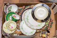 Top-down view of a cardboard box filled with assorted chinaware including cups, plates, bowls, and two figurines. Visible cracks on some plates.