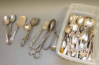 Overview of various vintage silver-toned serving utensils including large spoons, ladles, tongs, and pie servers on a flat surface with some utensils grouped next to a plastic container.