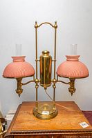 Full front view showing the brass body, two pink ribbed milk glass shades with clear glass chimneys on top, positioned on a wooden surface.