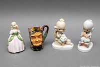 Four ceramic figurines with one pottery Toby mug. Includes a classic Royal Doulton Victorian lady figurine, Toby Mug, and three Mother & David’s children figurines.