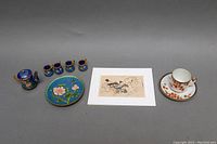 Photo showing entire lot with cloisonné miniature tea set, porcelain teacup and saucer, and Asian print laid out on grey surface