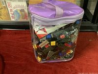 Photo showing large clear bag with purple trim and handle filled with assorted Lego pieces in various colors and shapes on a red surface.