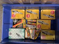 Blue bin containing multiple yellow NGK spark plug boxes and individual spark plugs inside smaller boxes and loose.