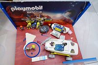 Playmobil Space Exhibition Box set components including spaceship, figurines, rock model, and space accessories arranged in front of the backdrop box.