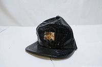 Front view of the black croc-embossed snapback hat with a gold studded metal plate accent.