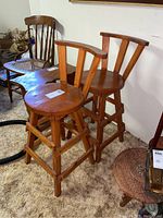 Pair of mid-tone brown wooden bar stools with round seats and three vertical slats forming backrest. Four legs with cross braces. Carpeted floor background.