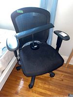 Front angle view of the black mesh office chair with padded seat, visible caster wheels, and part of black desk lamp on the right side. Light blue curtain background.