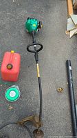 Full view of gas trimmer laid on asphalt with red jerry can, coil of green trimmer line, and two black poles/tools alongside.