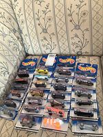 Photo of 25 Hot Wheels die-cast cars in original sealed packaging displayed on a floral patterned couch, showing a variety of models packed in blister card format with Hot Wheels blue backing.