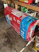 Image showing large unopened bag of Roxul ComfortBatt insulation with red and blue packaging, placed on a dirt floor near wooden shelves.