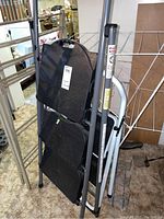 Photo showing the aluminum step ladder with black steps and white two step ladder next to wire clothes drying rack in area with other household items.