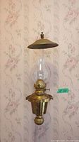 Full front view of one brass wall hanging oil lamp with bulbous glass chimney and rounded brass canopy against floral wallpaper.