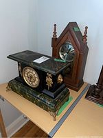 Photo showing two wood mantel clocks on a table, the front one with green marble-like finish, weathered dial, brass lion head detail, and brass feet. The back clock with a triangular wooden case and silver dial.