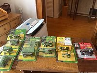 Overview of nine John Deere scale model tractors and farming accessories, all in original plastic blister packaging on bright green and yellow cardboard backing with some splits in packaging visible.