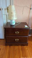 Front view of the wooden nightstand with two drawers topped by a white ceramic lamp with floral shade.