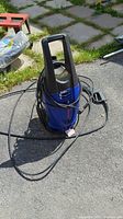 Side view of the blue and black Simoniz 1500 pressure washer unit with hose coiled around it and attached power cord, set on pavement outdoors.