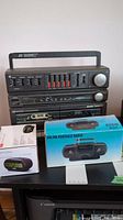 Three radios on table: RCA alarm clock radio in box, AM/FM boom box radio, and portable Bass Boost cassette deck radio.