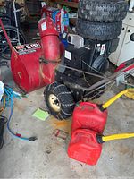 Photo showing the red snow blower with yellow handle, one tire with chains mounted and two extra tires stacked behind, two red gas tanks in front
