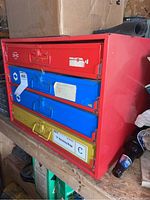 Front angled view of metal tool box showing red frame and three drawers in red, blue, yellow with handles and labels.
