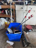 Photo of a blue bin filled with vintage hand drills, clamps, and garden tools with a folding two wheeled cart behind it, also visible is a small metal toolbox on the side.