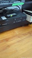 Photo of black Magnavox VCR DVD combo unit on a wooden floor shelf, with remote control on top and some cables behind.