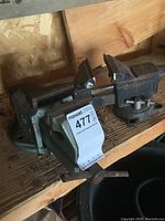 Photo of two heavy duty metal vices placed on a wooden shelf. One vice has a Maxsold tag attached showing lot number 477.