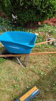 Side view of blue plastic Yardworks wheelbarrow with wooden handles and single black tire. Positioned on grass with garden background.