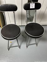 Two black folding chairs with round seats and rectangular backs, metal frames, standing side by side.