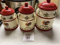 Three ceramic canisters for coffee, tea, and sugar with red lids and rooster/floral painted decoration on a white background