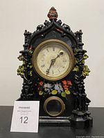 Front view of antique American clock showing black painted metal front with floral decoration and round clock face with Roman numerals.