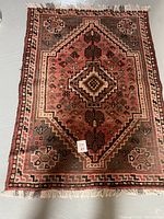 Full view of the antique wool rug showing its geometric and floral design in red, brown, cream, and black tones with fringed edges.