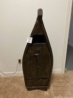 Full view of solid wood cabinet shaped like a boat with pointed top, carved details on door and drawer, standing on carpeted floor.