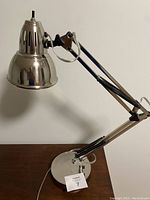 Desk lamp with stainless steel finish, adjustable arm, and clamp base shown on wooden surface against white wall.