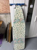 Full view of the ironing board with floral fabric cover hanging vertically against a white door with a mirrored wardrobe beside it.