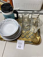 Photo showing a rectangular ornate metal tray holding six white ceramic plates with blue floral borders, six glass drinking vessels including mugs and glasses, and a blue insulated travel coffee cup with handle and lid.
