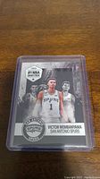 Image of a Victor Wembanyama basketball rookie card from 2023 featuring San Antonio Spurs in protective sleeve on wooden surface.