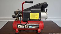 Side view of Clarke air compressor showing the red tank with Clarke Air logo, motor housing, handle and pressure controls.