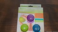 Photo showing the Restore Hand Therapy Kit packaging with the soft purple, medium green, and firm blue therapy balls visible alongside the exercise guide.
