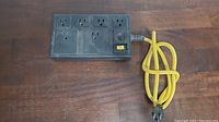 Full view of the black power bar with five outlets and yellow cord on wooden surface