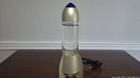 Front view of the rocket ship lava lamp showing gold base, clear liquid chamber with blue and silver particles, and blue pointed top.