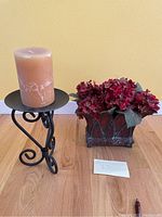Photo of red silk floral arrangement in a decorative dark metal planter and large metal candle stand holding a pillar candle on a wood floor against a yellow wall.