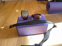 Image shows Garmin Forerunner 305 watch, Polar heart monitor device, and adjustable black heart monitor strap laid on a yoga block on a hardwood floor.