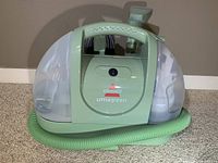 Front and side view of the Bissell Little Green carpet cleaner showing water tanks, handle, and hose attached at front.