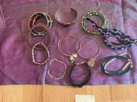 12 assorted bracelets of different styles including rhinestones, leather, pearl beads, braided and metal bangles, arranged on a purple cloth background.