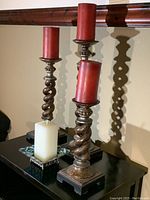 Three spiral candlesticks with square bases on black surface, holding tall red pillar candles and a white square candle on metal stand.