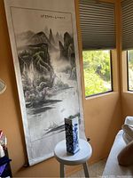 Photo of vertical Chinese silk watercolor painting showing mountain landscape and calligraphy with blue and white porcelain vase in front on small white table near window.
