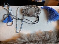 Photo of three jewelry items: a long blue rhinestone necklace with tassels, a silver beaded necklace, and a silver bracelet on a light fabric surface.