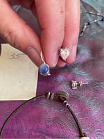 Close-up hand holding two earrings: one blue moonstone and one heart-shaped cubic zirconia, with braided leather bracelet below.