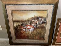 Framed Italian village landscape painting showing hillside houses with red roofs and a distant tower under a warm sky