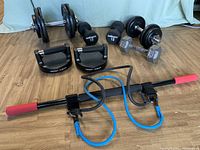 Photo showing multiple dumbbells, push-up handles, and resistance band rod on wooden floor with cloth backdrop.